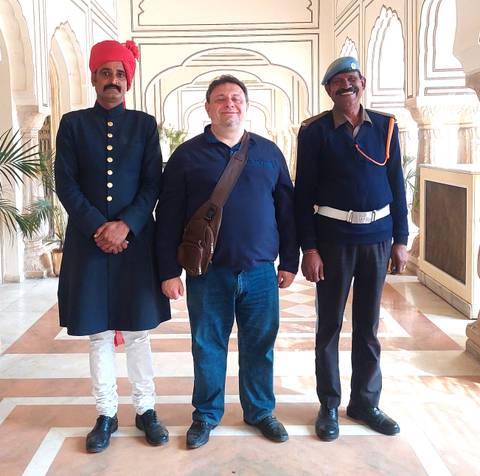       Tourist poses between hotel doorman and security guard inside ornate lobby.
  