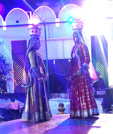       Blurred stage shot of two traditionally dressed dancers conversing under blue stage lights.
  