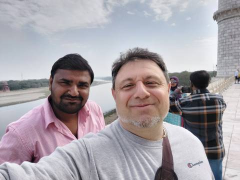       Casual outdoor selfie of two men smiling by a riverside monument with other tourists behind.
  