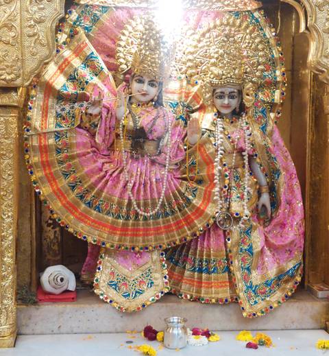       Ornate pink-clad Hindu deities adorned with jewelry and garlands inside gilded temple shrine.
  