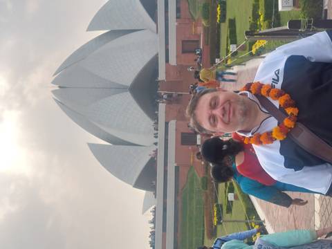       Man wearing a marigold garland posing in front of the Lotus Temple with other visitors in the background
  