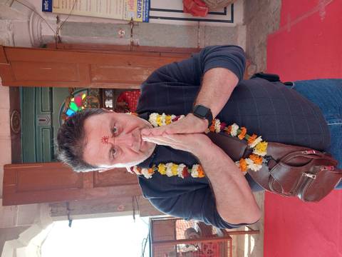       Man with garland and prayer gesture inside a historic doorway with stained glass above
  