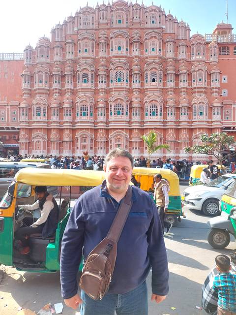       Man posing in front of the busy Hawa Mahal façade with tuk-tuks and crowds on the street
  