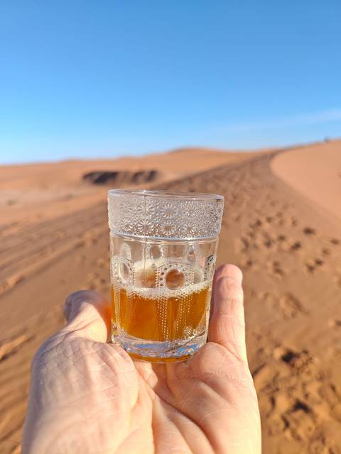       Hand holding a decorated glass of tea against sweeping Sahara sand dunes at golden light
  