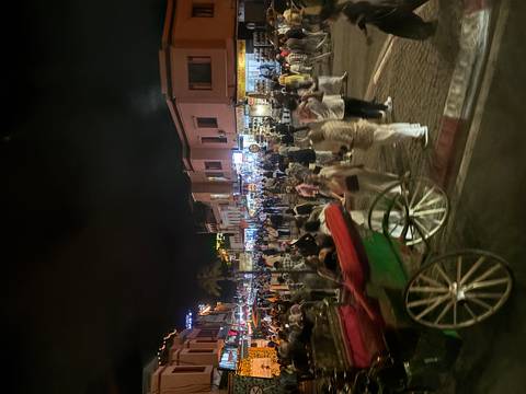       Night market in Marrakech teems with people, stalls and carriages under neon lights.
  