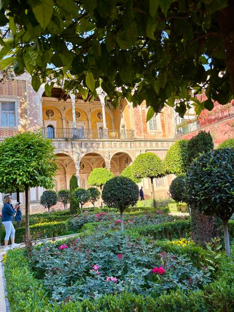       Lush garden courtyard with trimmed trees and arches of a yellow-walled palace, with a visitor taking photos.
  