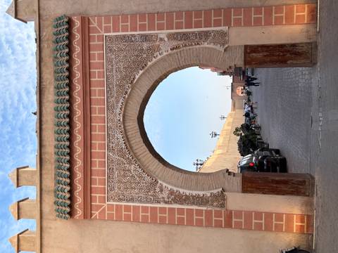       Tall decorative city gate with intricate plasterwork framing a view of a cobblestone street and blue sky.
  