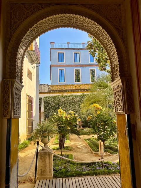       View through a carved Moorish archway onto a small flower-filled courtyard and pale blue villa.
  