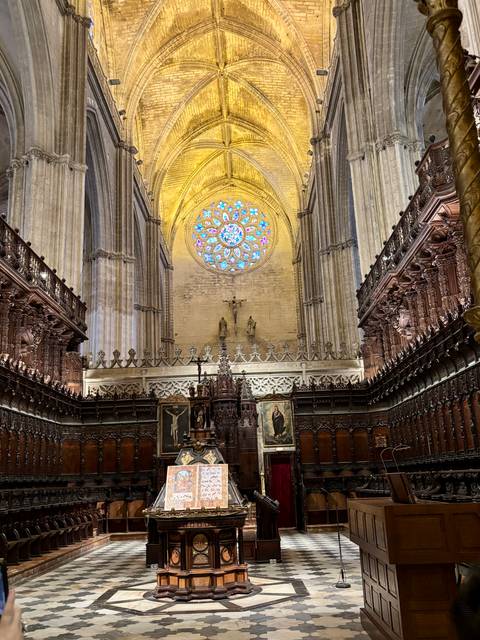       Interior of a grand gothic cathedral choir with soaring columns and a colorful rose window above the altar.
  