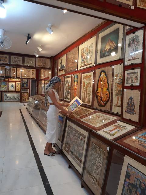       A woman in a white dress browses colorful papyrus art inside an Egyptian gallery.
  