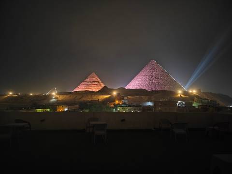       Illuminated pyramids of Giza glowing at night, seen from a rooftop with city lights and a spotlight beaming into the sky.
  