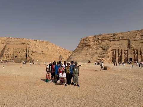       Tour group stands in the vast sandy forecourt of Abu Simbel temples under a hazy sky.
  