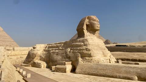       The Great Sphinx of Giza in the desert with pyramids and hazy sky behind.
  