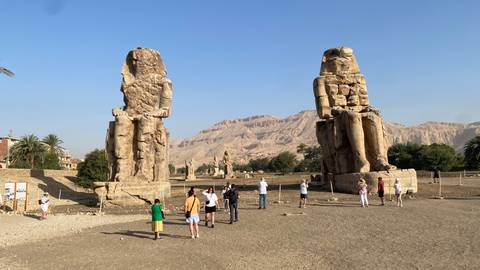       Visitors explore the colossal seated statues known as the Colossi of Memnon against a mountainous backdrop.
  
