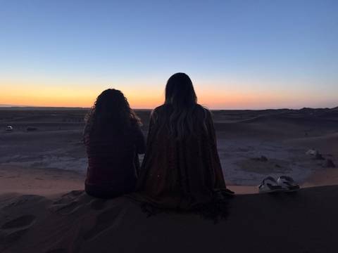       Two travellers sit on a desert dune watching the colourful glow of sunset spread across the horizon.
  