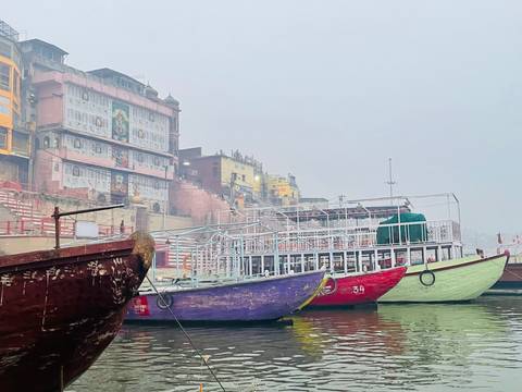       Colourful wooden riverboats sit moored along a hazy ghatside lined with painted buildings and murals.
  