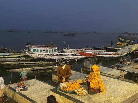      Vendors arrange flower offerings beside the dark river as boats float in the early dawn gloom.
  