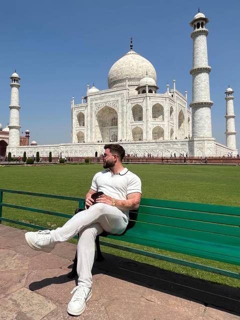       Traveller relaxes on a bench with the gleaming white marble Taj Mahal rising behind him.
  