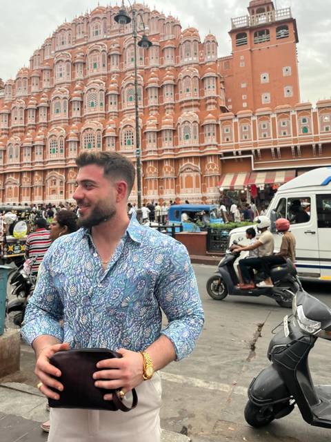       Man smiles in front of the ornate pink façade of Jaipur’s Hawa Mahal amid busy street life.
  