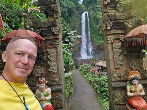       Selfie at ornate Balinese gate framing tall rainforest waterfall.
  