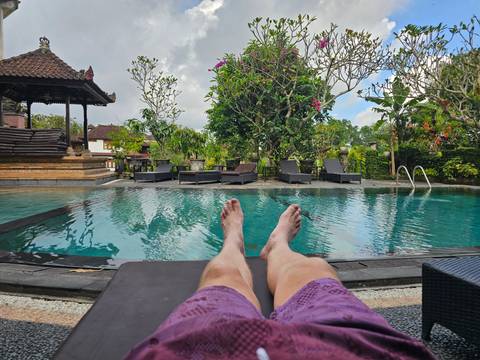       Person relaxing on lounge chair with feet toward serene pool and tropical garden.
  