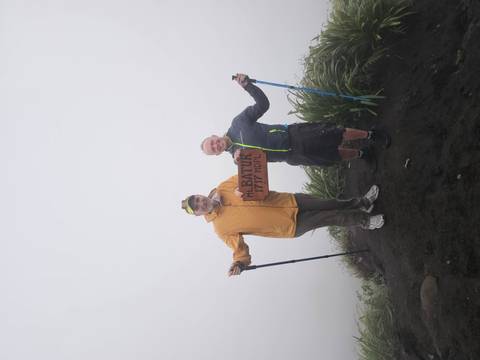       Two hikers at foggy Mount Batur summit holding wooden altitude sign.
  