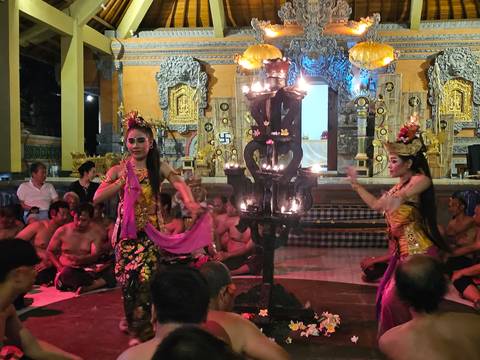       Balinese dancers performing a traditional temple dance surrounded by seated male chanters and ornate golden carvings, lit by a tall candelabra.
  
