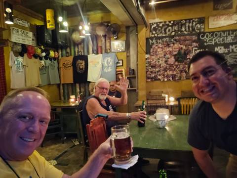       Three men raising beers and smiling in a cozy, dimly lit bar decorated with t-shirts and handwritten chalkboard signs.
  
