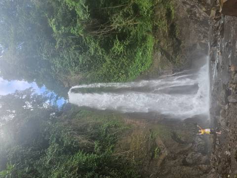       Tall waterfall plunging down a lush cliff into a rocky pool with a lone person in a yellow shirt posing at the base.
  