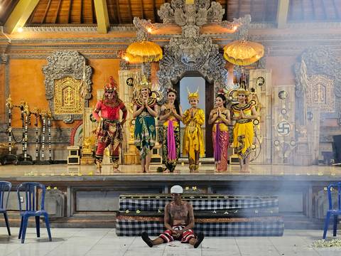      Colorful Balinese dance troupe posing on an ornate temple stage decorated with carved stonework and golden umbrellas.
  