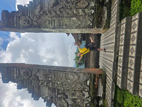       Traveler playfully balancing on one leg beneath a massive stone Balinese gateway with patterned tile steps and cloudy skies above.
  