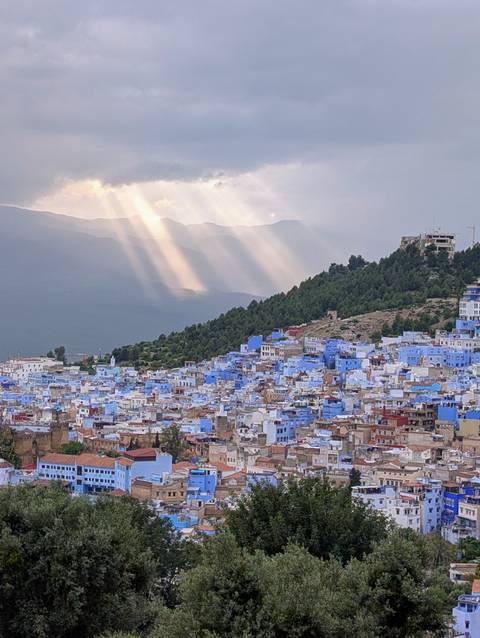       Chefchaouen’s blue-painted houses sprawl across a hillside as sunbeams pierce clouds above the Rif Mountains.
  