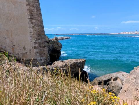      Turquoise Atlantic waves crash against rugged rocks and an old stone fort wall under a bright sky.
  