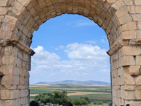       Stone Roman arch at the ruins of Volubilis frames rolling farmland and distant mountains under a blue sky.
  