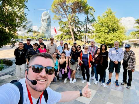       Cheerful selfie of a tour group in a modern city park with a distinctive curving skyscraper in the background.
  