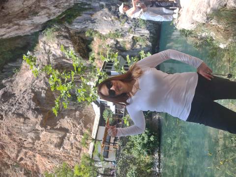       Woman posing beside a turquoise spring and cliffside Tekke house at Blagaj.
  