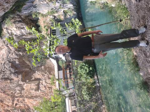       Man posing beside clear spring water and a cliffside restaurant similar to Blagaj’s setting.
  