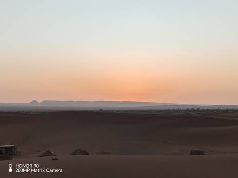       Subtle orange glow of sunrise over distant desert ridges and dark foreground dunes.
  