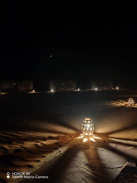       Nighttime desert camp with lanterns illuminating sand ripples and simple tents in darkness.
  