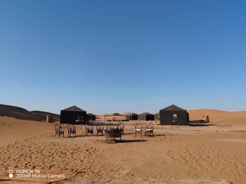       Fixed desert camp with black canvas tents and metal fire pit under a clear blue sky.
  