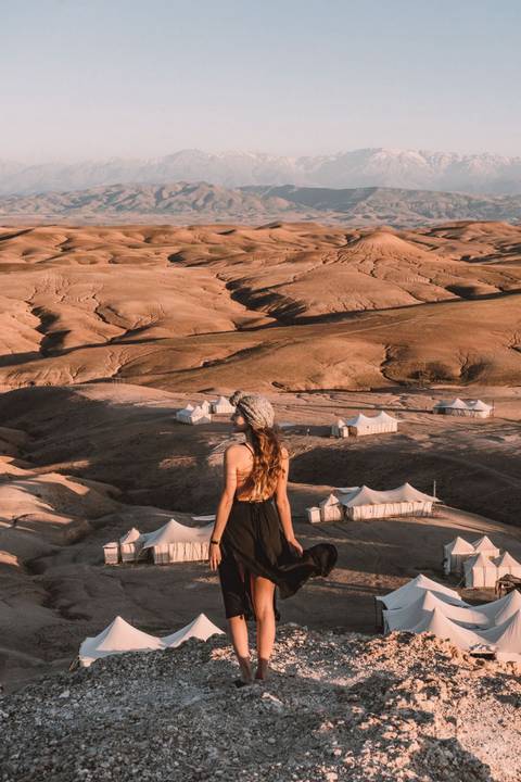       Young woman gazes over remote desert camp of white canvas tents scattered across rolling tan hills.
  