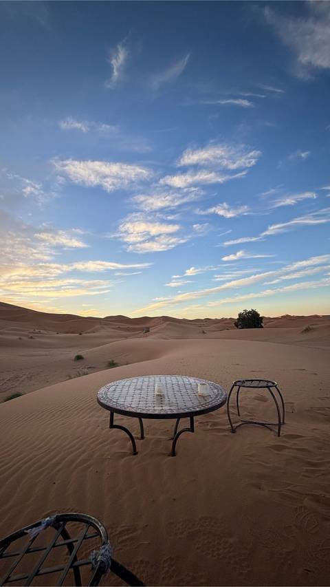       Soft evening light over rolling sand dunes beneath a pastel sky; low desert camp seating visible in foreground.
  