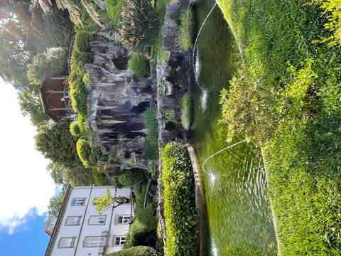       Lush garden with artificial grotto, pond and spraying fountain beside a stately home.
  