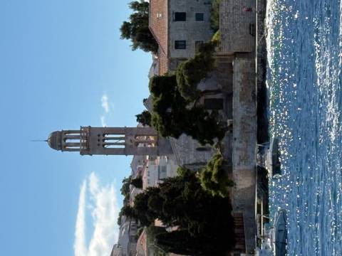       Stone church and bell tower on a coastal promontory viewed across sparkling blue sea.
  