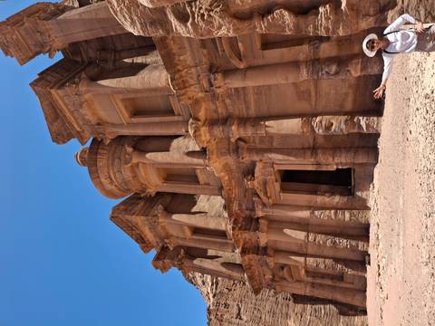       Sandstone façade of Petra Monastery towering above visitor’s outstretched arm.
  