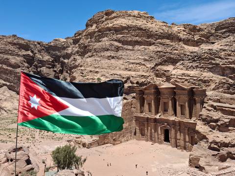       Jordanian flag waves in foreground overlooking majestic Petra Monastery and surrounding cliffs.
  