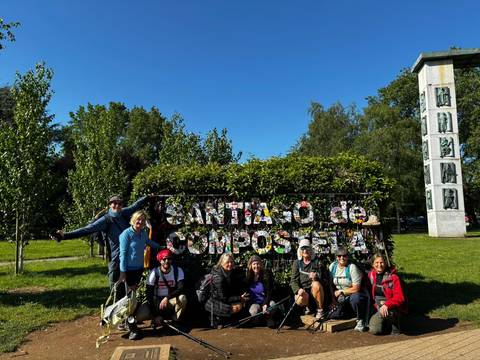       Pilgrim group poses by leafy Santiago de Compostela sign in sunny park.
  