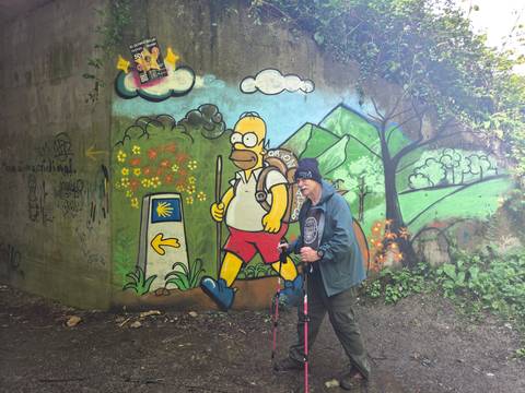       Pilgrim with walking sticks stands by colourful Homer Simpson Camino mural under bridge.
  