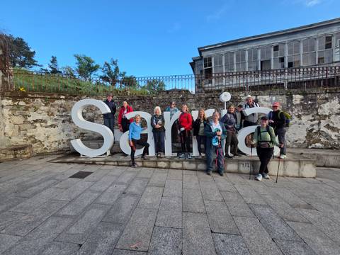       Large walking group gathers around white Sarria town sign on paved square.
  