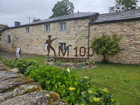       Sign for KM100 Casa do Rego on the Camino set in lush garden beside stone wall.
  